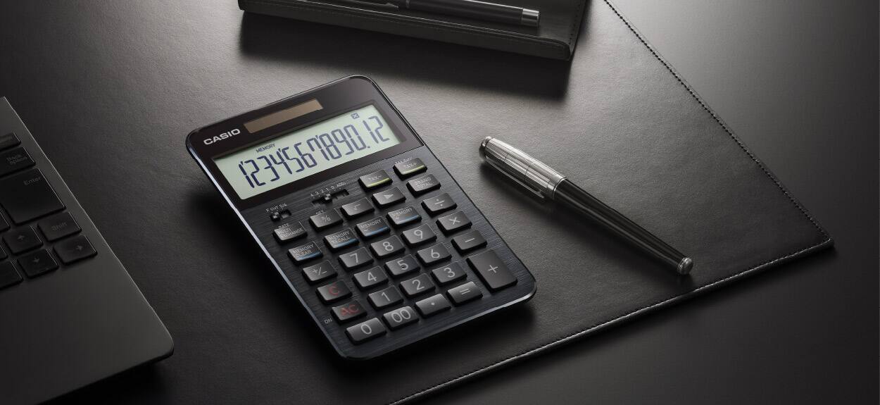 Der schwarze Premium-Rechner von CASIO auf einer Schreibtischunterlage neben einem Premium-Kugelschreiber und einem Laptop