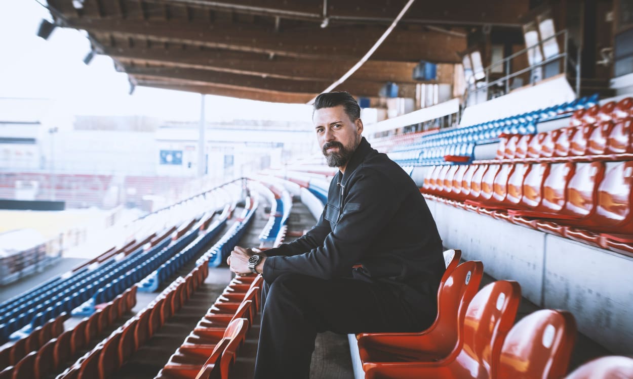 Sandro Wagner sitzt auf roten Plastikstühlen einer Tribüne im Fußballstadion