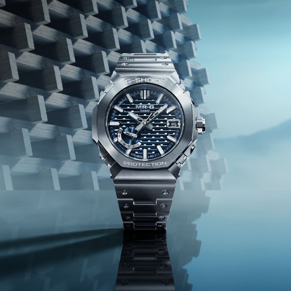 Die silberne G-Shock MRG-B2100D-2A Armbanduhr mit Metallarmband vor einem geometrisch strukturierten Hintergrund.