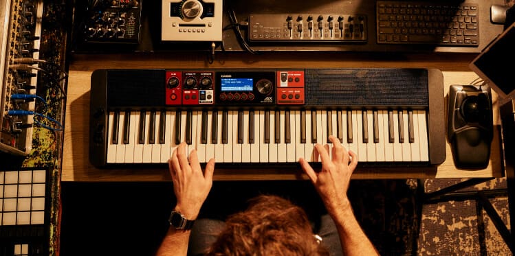 Blick von oben auf Hände, die auf einem Digitalpiano spielen, das auf einem Tisch inmitten von Tontechnikgeräten steht. Das Bild verlinkt zur Übersichtsseite: Vokalsynthese