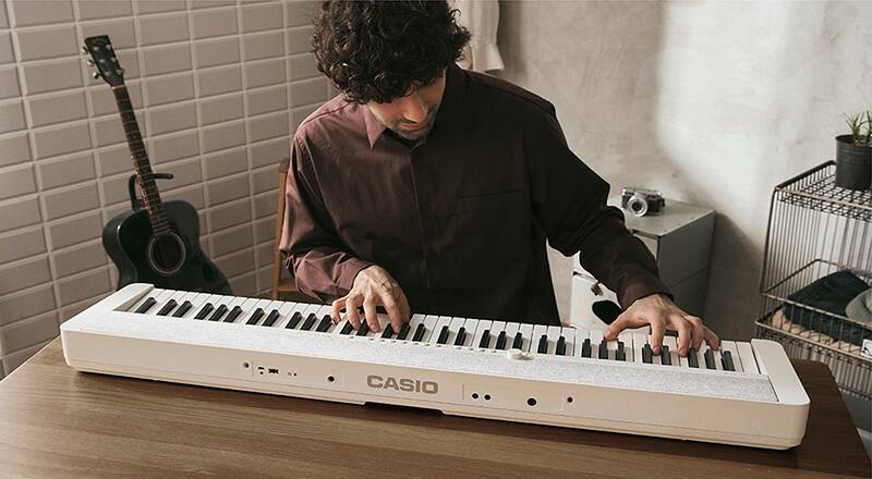Ein Mann sitzt an einem Holztisch und spielt auf dem weißen Casiotone CT-S1-76 Digitalpiano
