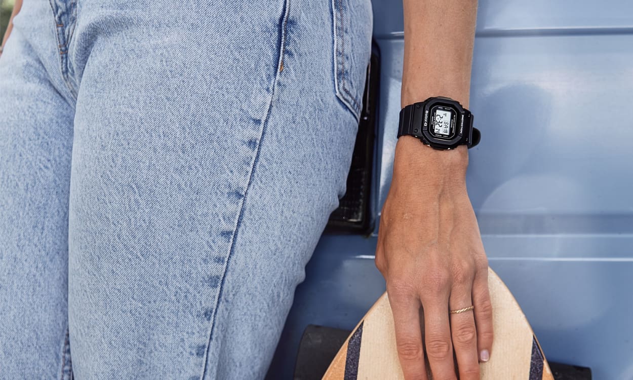 Eine Skaterin trägt die Baby-G BGD-560 1ER mit ihrem Skateboard und lehnt sich an einen Van.