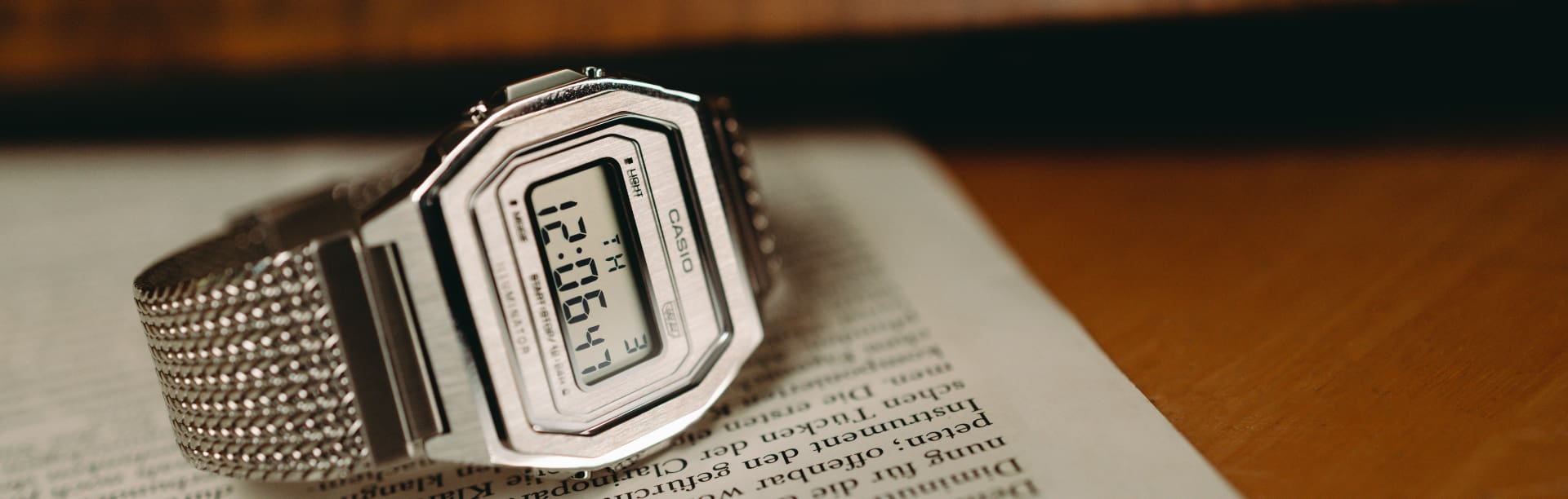 Die CASIO Vintage Collection Uhr A1000MA-7EF mit eckiger, digitaler Anzeige und silberfarbenem, kleingliedrigem Armband liegt auf einer Zeitschrift