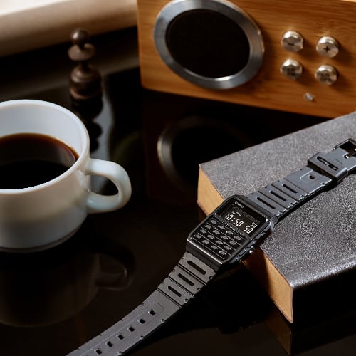 Casio CA-53WF-1B in schwarz liegend auf einem Buch neben einer Tasse Kaffee auf einem Glastisch