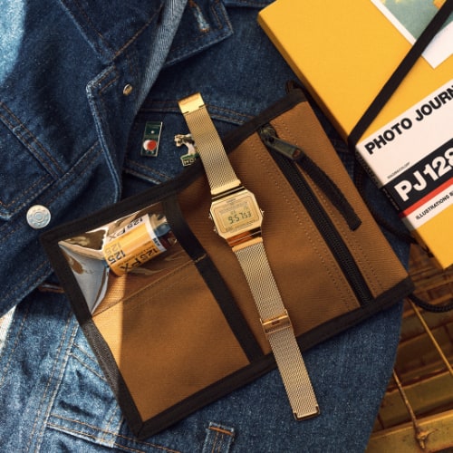 Casio Vintage A700 in gold mit Milanaise-Armband auf einem Portemonnaie neben einer Jeansjacke und einen Kamerafilm.