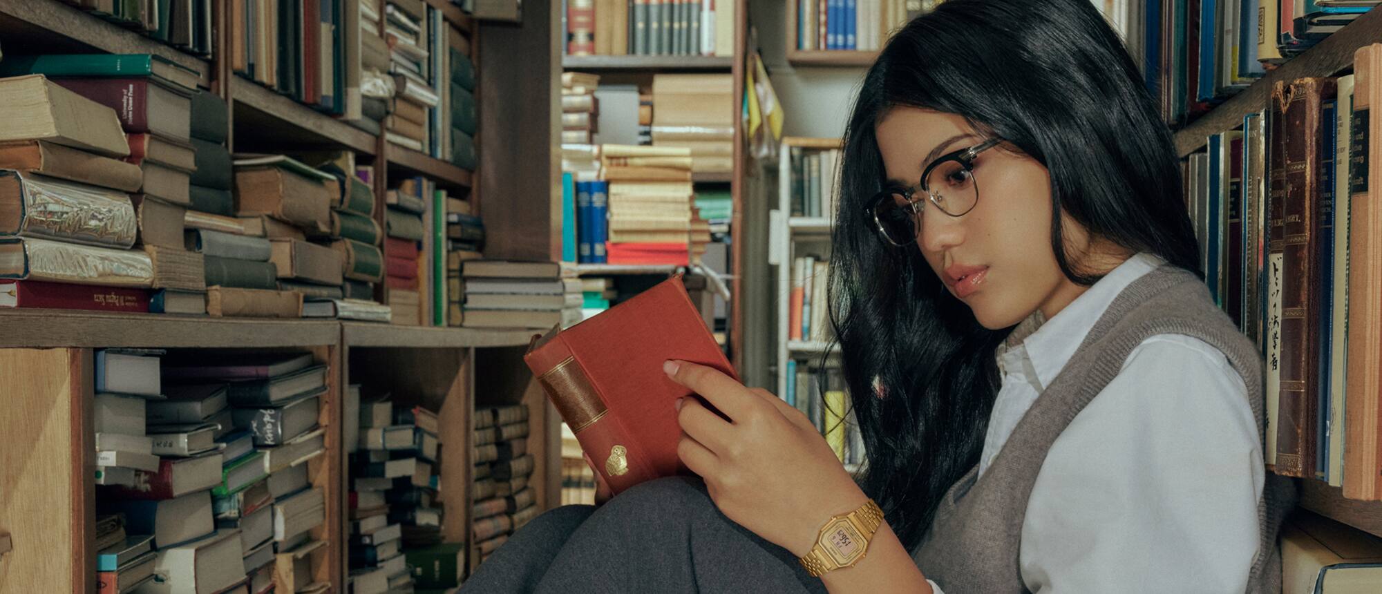 Eine Frau mit braunem Haar und Brille liest ein Buch in einer Bibliothek und trägt die CASIO Vintage Collection Uhr LA680WGA-9.