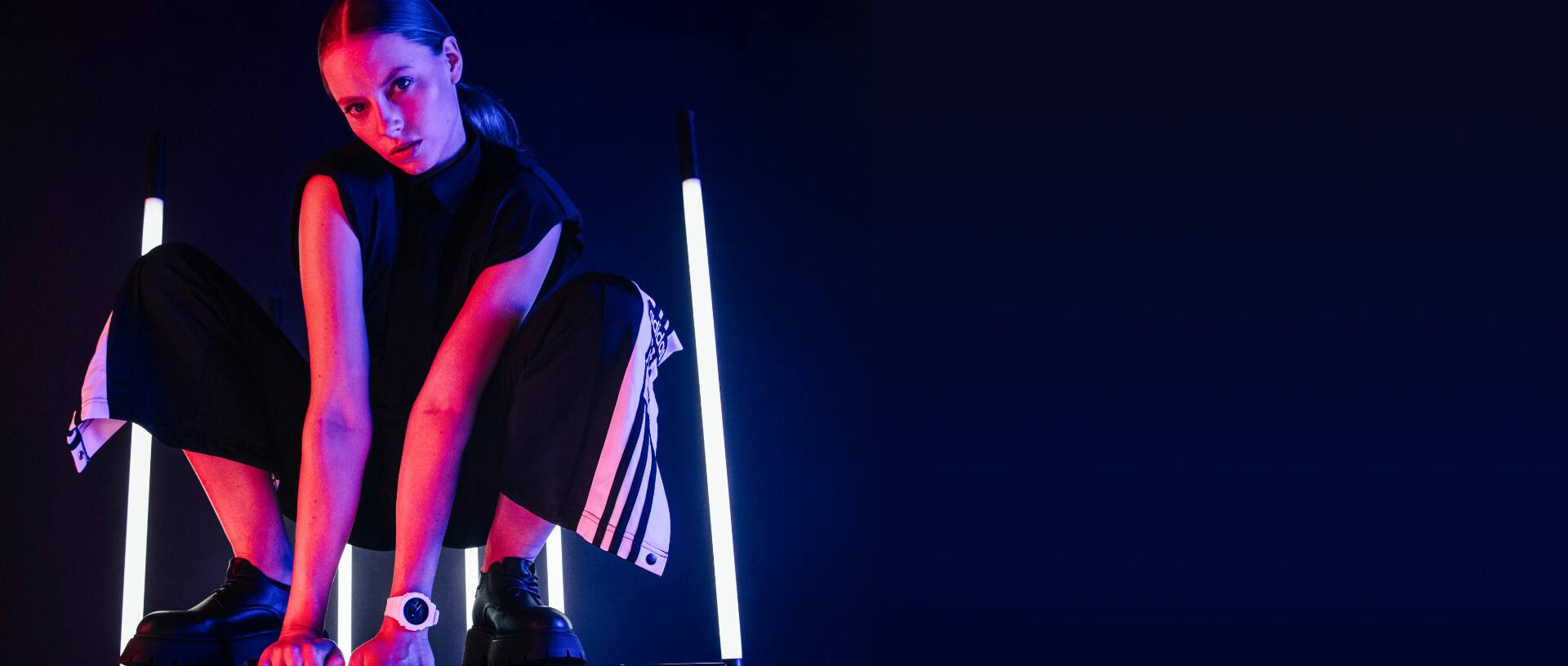 Die weiße GA-2100 Armbanduhr mit analoger Anzeige am Handgelenk einer Frau in schwarzer Kleidung. Im Hintergrund sind lila und pinke Neonröhren zu sehen. Das Bild verlinkt zur Produktseite. 