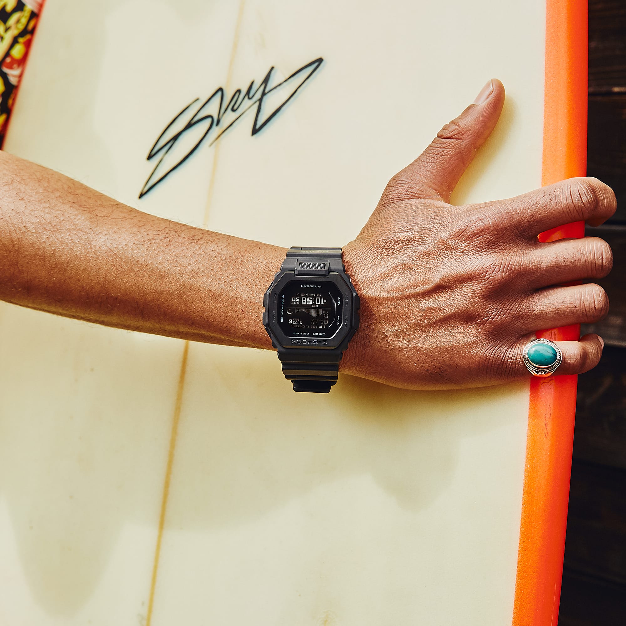 Jemand trägt ein Surfboard, am Handgelenk trägt die Person eine G-Shock GBX-100NS-1 Armbanduhr mit Digitalanzeige aus der G-SQUAD Reihe 