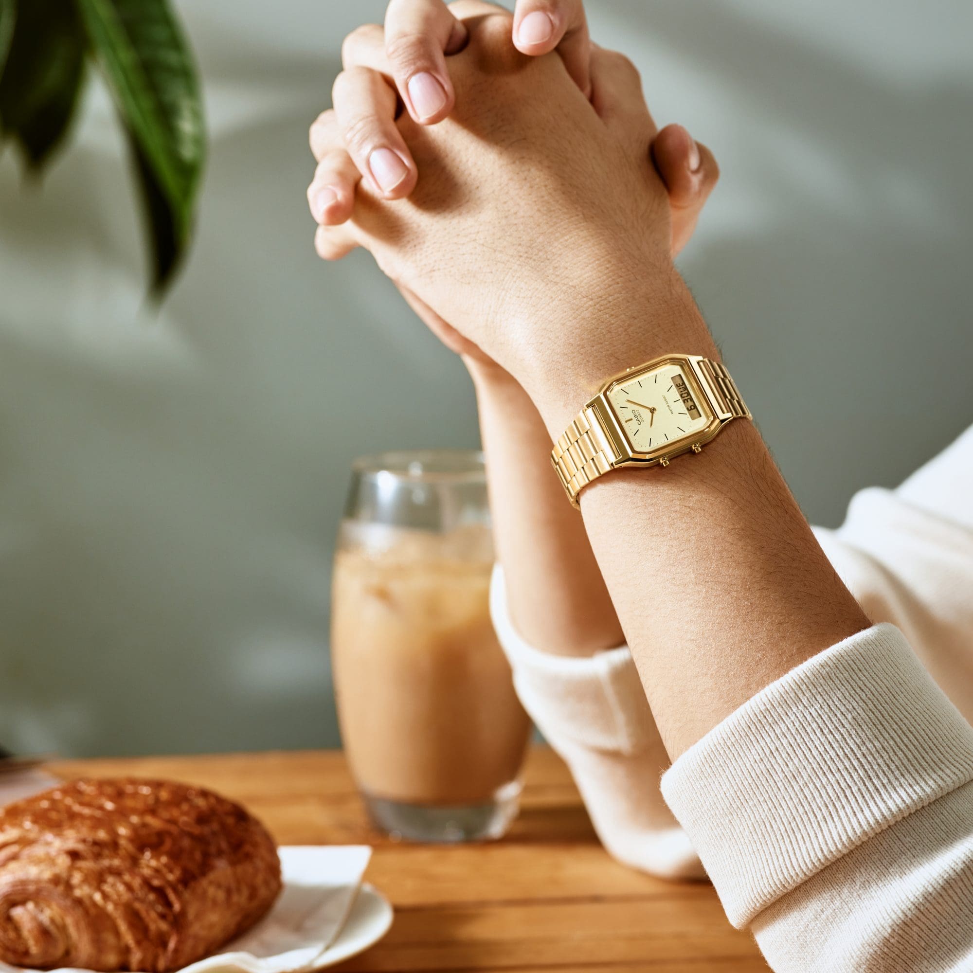Nahaufnahme eines Unterarms, die Ellenbögen sind auf einem Tisch abgelegt. Auf dem Tisch liegt eine Croissant und ein Iced Latte im Glas. Am Handgelenk der Person ist die goldfarbene CASIO Vintage-Uhr "AQ-230GA-9DMQ" zu sehen. Das analoge Zifferblatt ist beige gehalten, ganz unten ist eine kleine digitale Datumsanzeige.