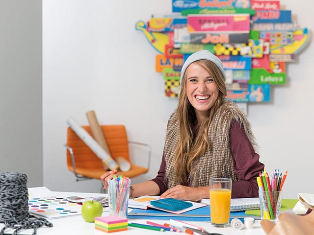 Eine junge Frau sitzt an einem kreativ gestalteten Schreibtisch mit Farbmusterkarten und Buntstiften. Auf dem Tisch liegt ein blauer Taschenrechner