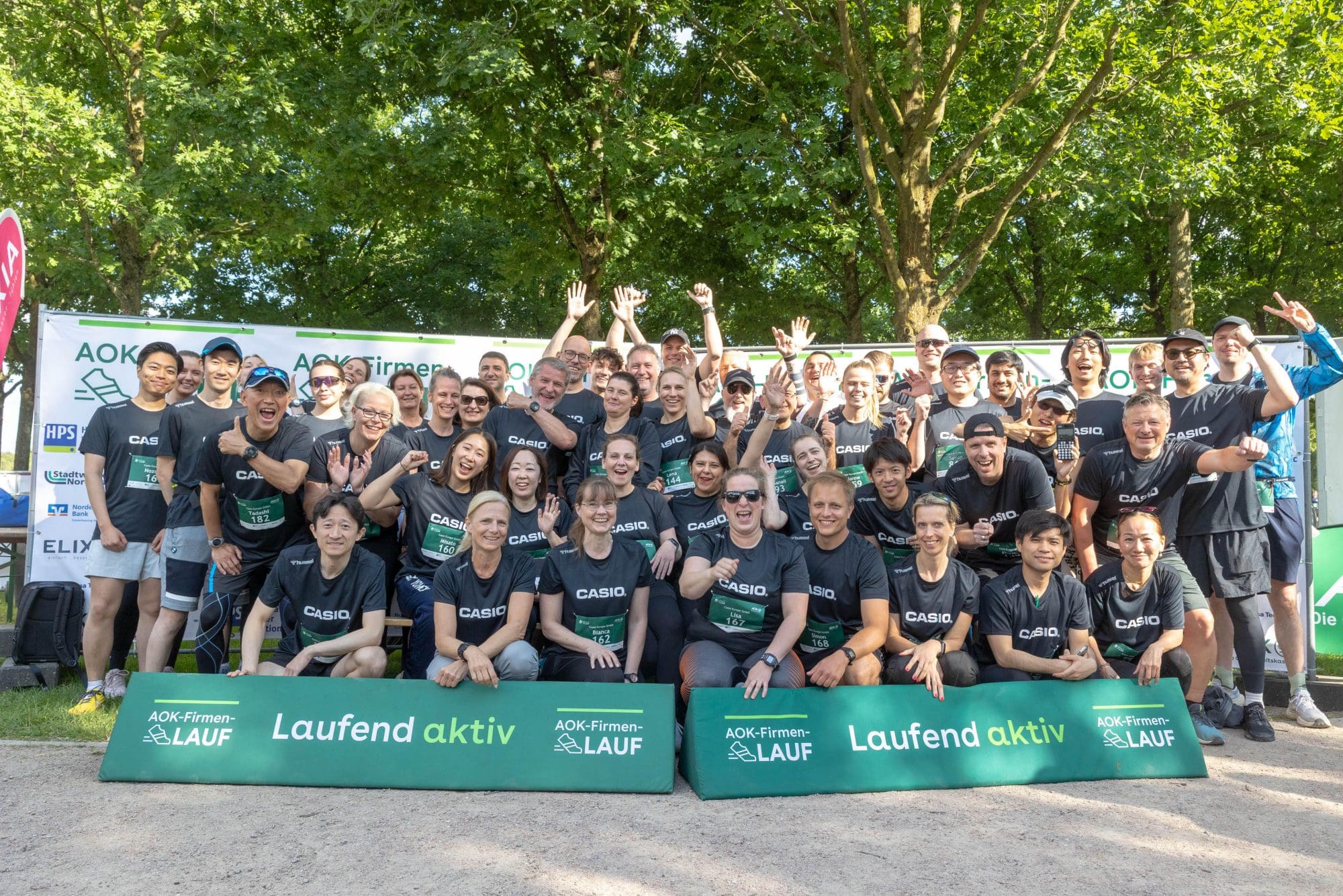 Casio Team beim AOK Firmenlauf Event in weißen Casio T Shirts vor Veranstaltungsbanner auf einer Wiese