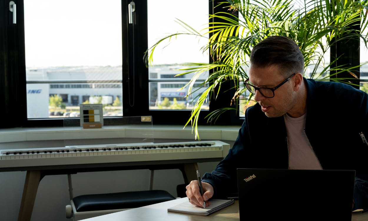 Ein Sales-Mitarbeiter sitzt am Schreibtisch und notiert etwas im Notizbuch. Im Hintergrund ist ein CASIO Keyboard zu sehen