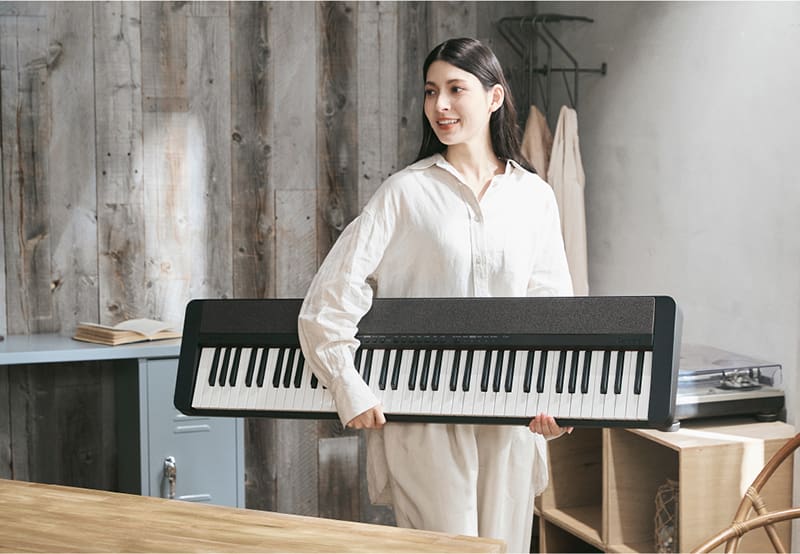 Eine hell gekleidete Frau trägt ein schwarzes CT-S1-76BK Digitalpiano unter einen Arm gehalten durch ein Büro