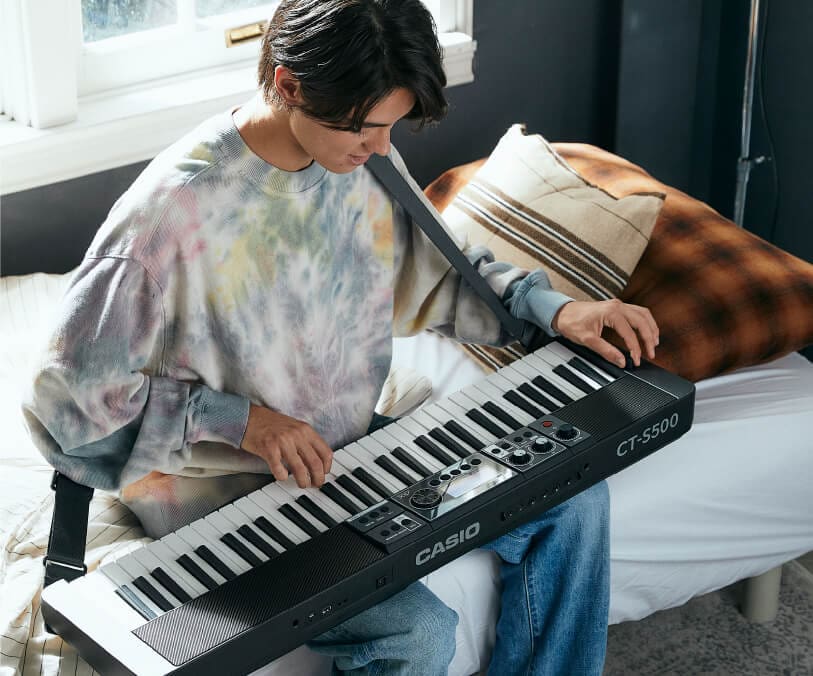 Ein junger Mann in einem hellen Batikpullover sitzt auf einem Bett und spielt auf einem Casiotone CT-S500, das auf seinem Schoß liegt