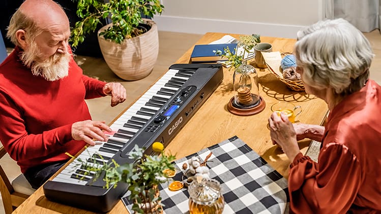 Ein alter Mann sitzt an einem Tisch und spielt auf dem Casiotone LK-S450 Digitalpiano, eine alte Dame sitzt ihm gegenüber und hört zu