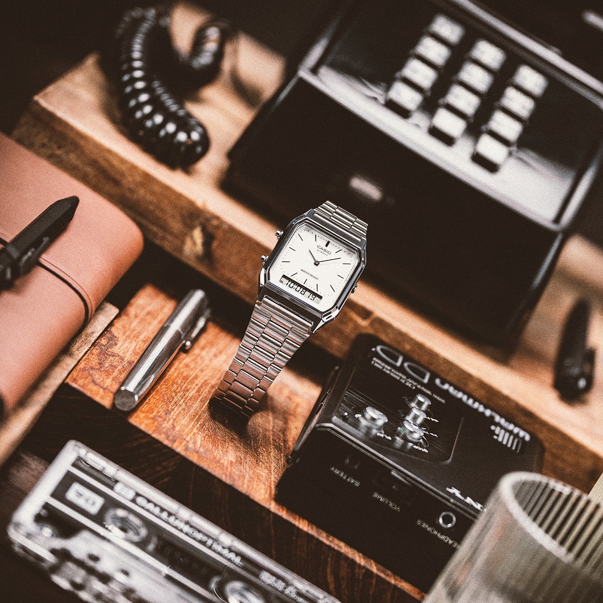 Das AQ-230 mit weißem Ziffernblatt und silbernem Armband auf einem Holztisch neben einem Notebook und einem alten Telefon