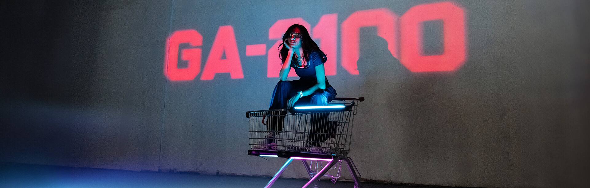 Wa22ermann sitzt auf einem Einkaufwagen, hinter ihr wird der Schriftzug GA-2100 in rot an eine Wand projiziert. Das Bild verlinkt zur Produktseite der GA-2100-7A7 Armbanduhr.