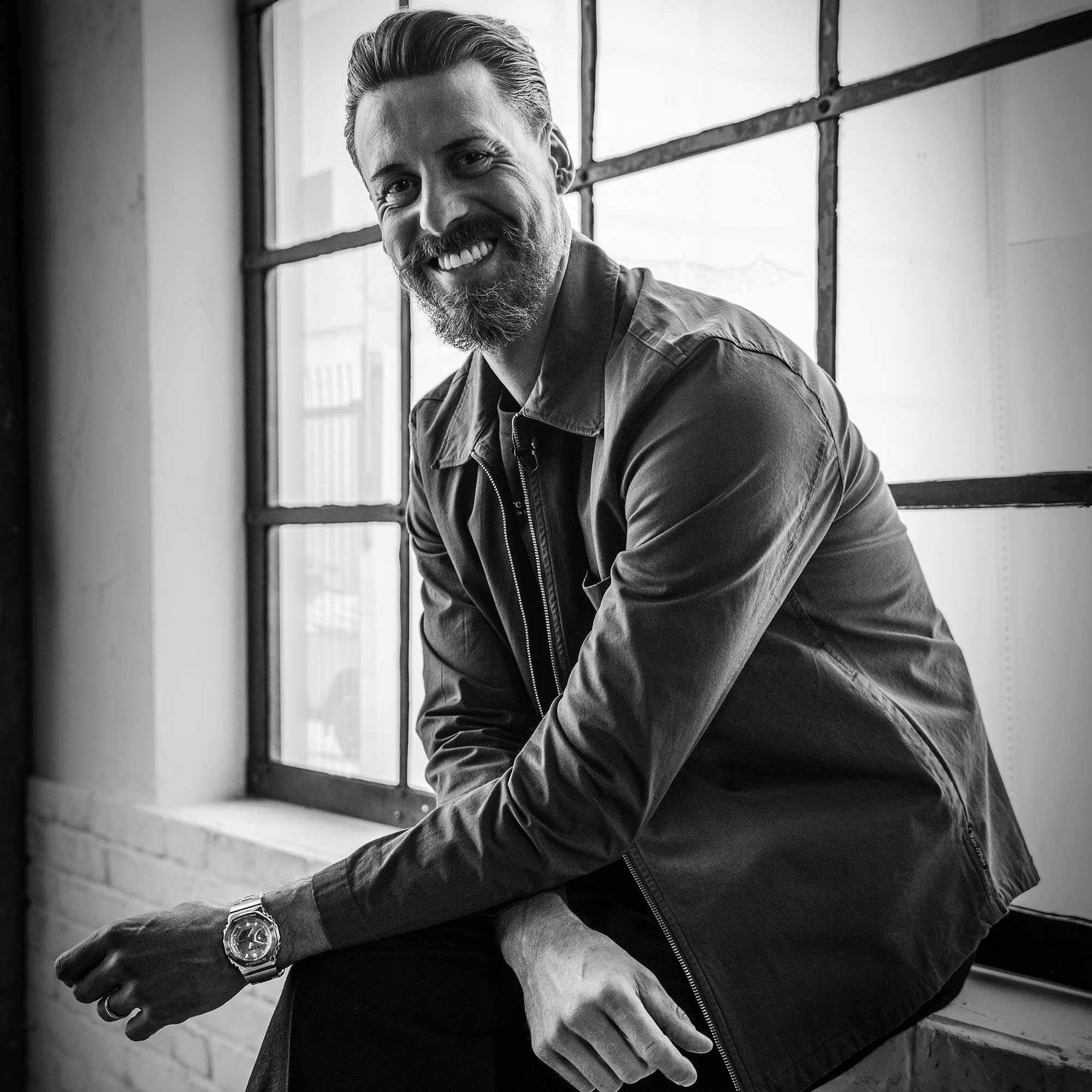 Trainer Sandro Wagner sitzt in Jeanskleidung auf einer Fensterbank und lächelt in die Kamera, dabei stützt er seine Arme auf den Knien ab.