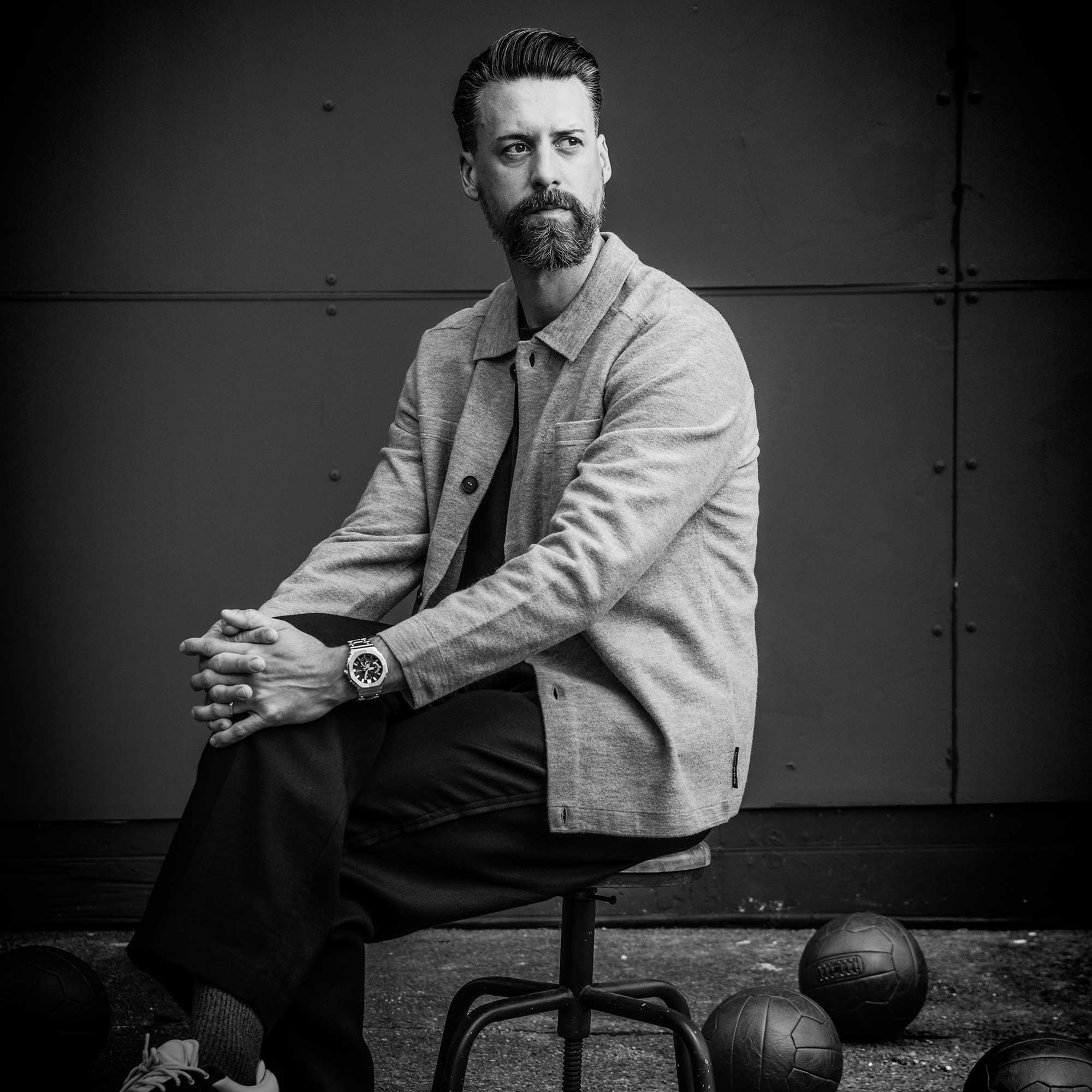 Schwarz-weiß Bild von Sandro Wagner auf einem Hocker, mit hellem Denim-Hemd und dunkler Hose. Am Handgelenk trägt der die Full-Metal Armbanduhr GMC-B2100 von G-SHOCK.