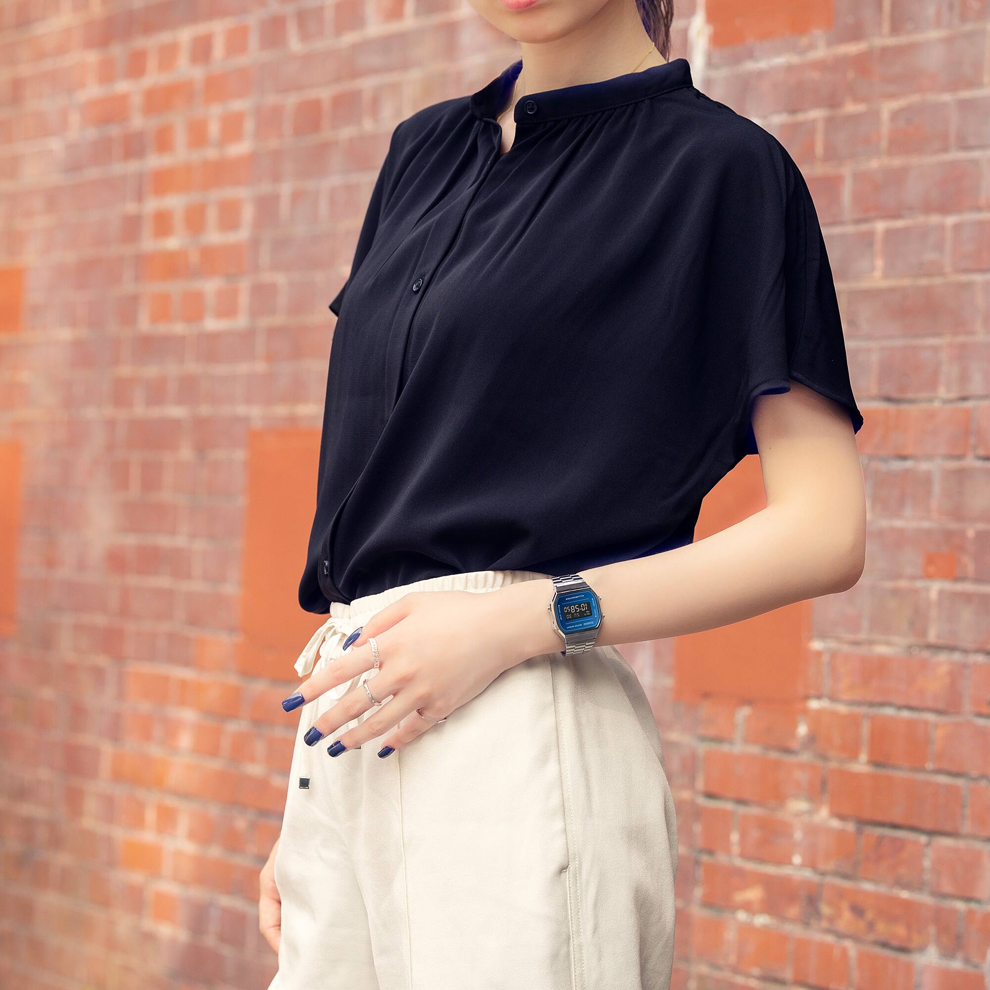 Ein Model steht vor einer Backsteinmauer in schwarzem Oberteil und beiger Hose. Am Handgelenk trägt sie die silberfarbene CASIO Vintage Uhr "A168WEM-2B" mit blauer digitaler Zeitanzeige.