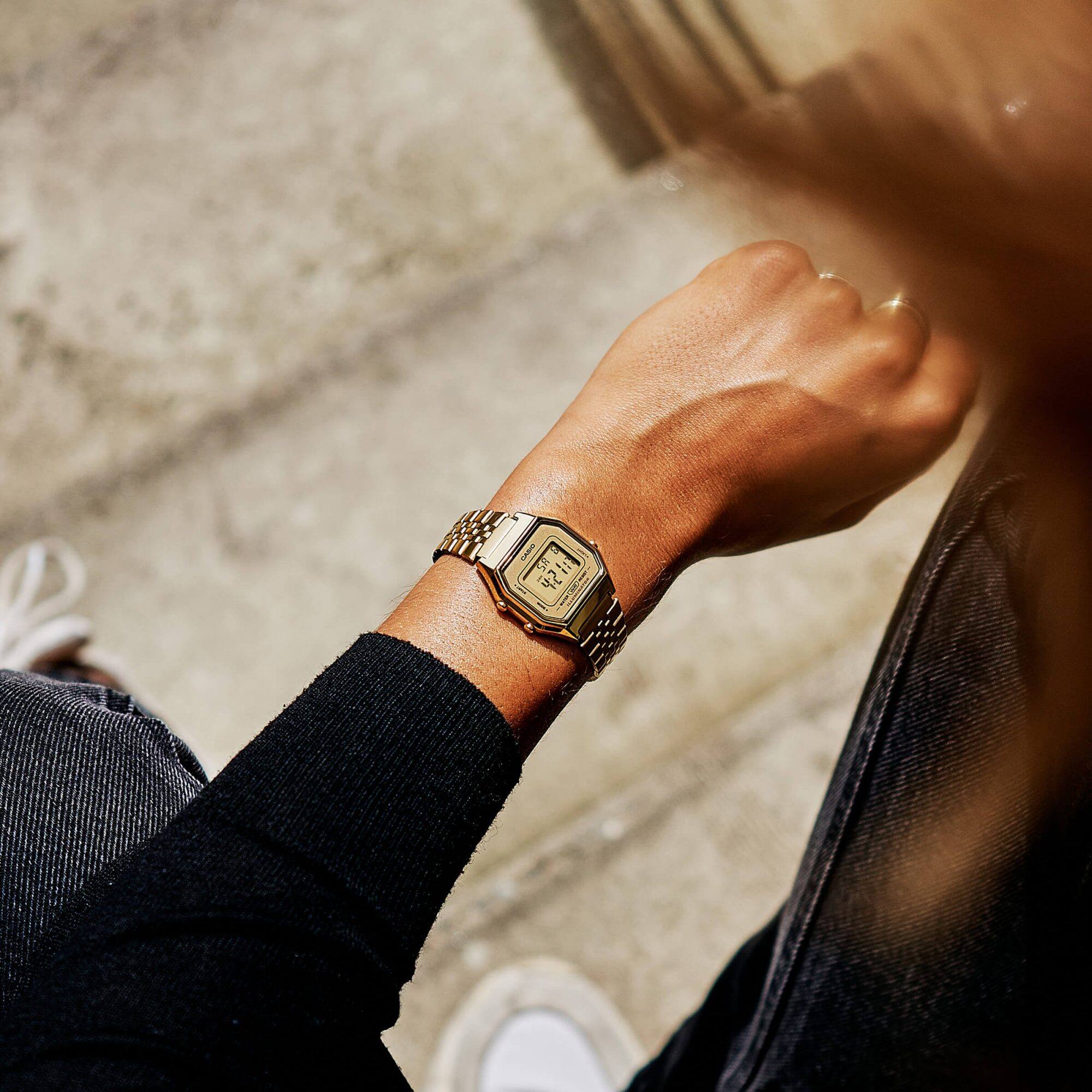 Aus der Vogelperspektive wird das Handgelenk einer Frau gezeigt, die die CASIO Vintage "LA680WEGA-9" Armbanduhr zeigt. Die Uhr hat ein goldfarbenes Armband und ein digitales Zifferblatt.
