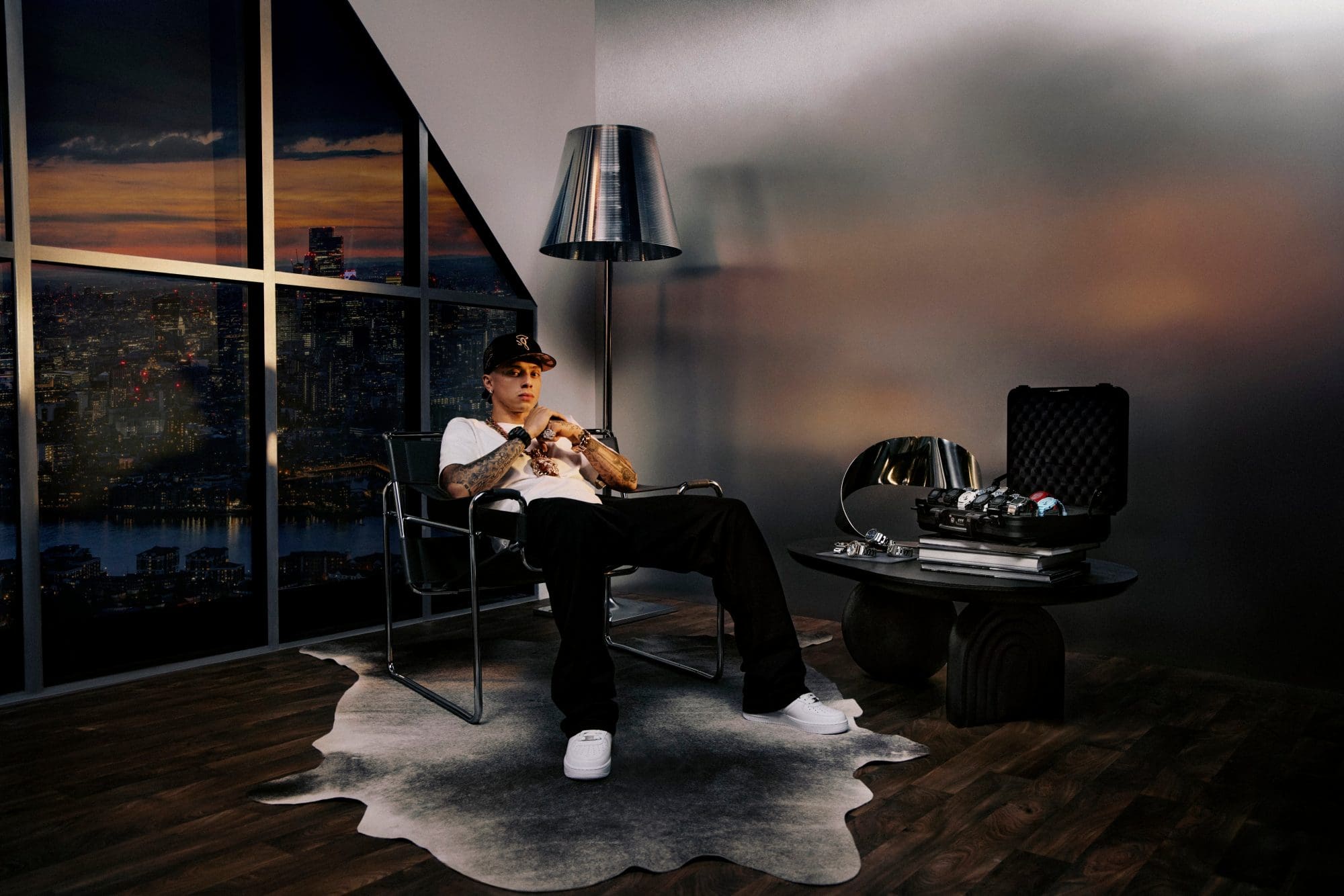 Central cee is chilling on a chair, wearing a G-SHOCK and has a suitcase full of watches on the desk next to him.