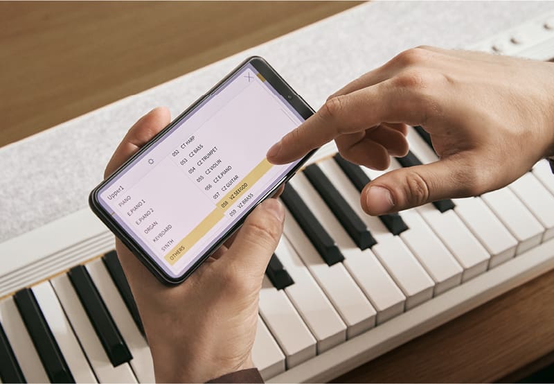 Vista desde arriba de un piano digital CT-S1-76 blanco y  una mano que sostiene un smartphone mientras la otra mano ajusta la configuración de sonido en la aplicación CASIO MUSIC SPACE