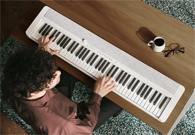 Un hombre sentado a una mesa de madera toca el piano digital blanco CT-S1-76WE