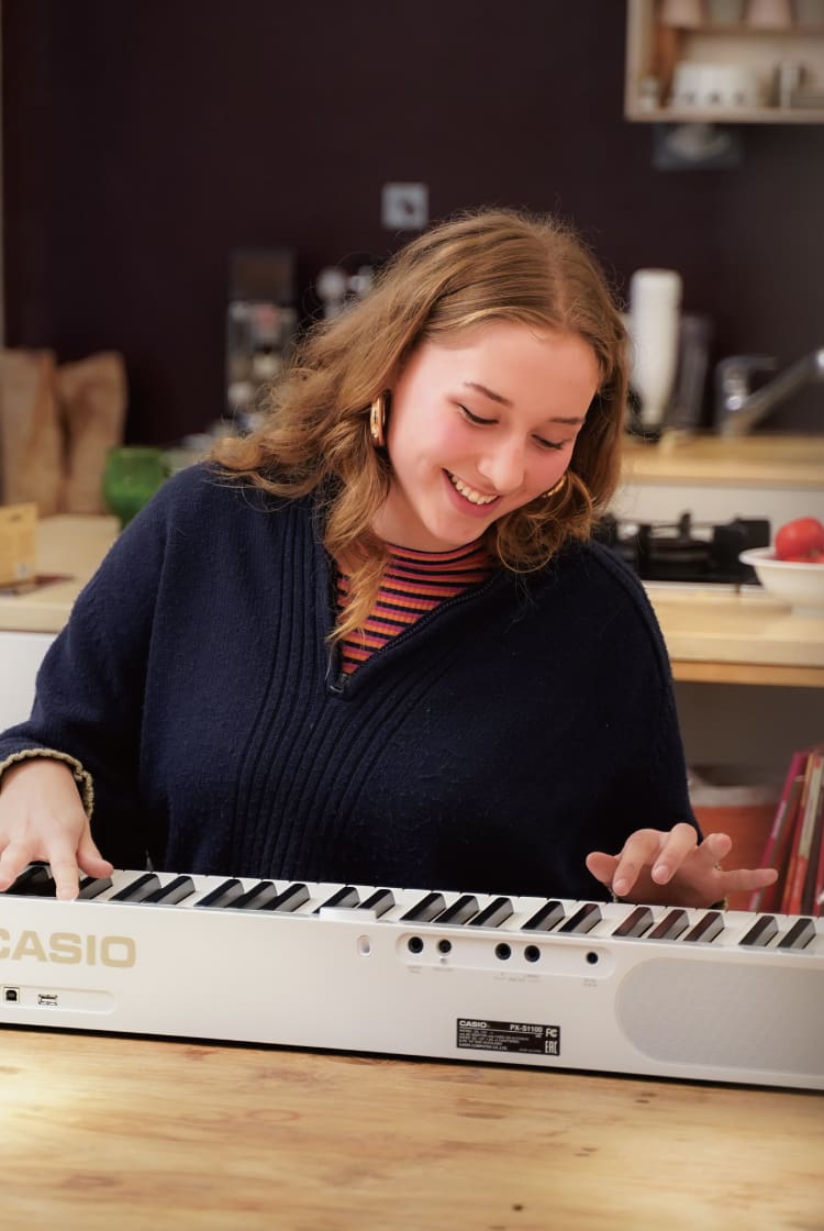 La estudiante Antonie Weber está sentada en la mesa de la cocina y toca su piano Privia blanco. La imagen enlaza con su página «La vida con Privia».