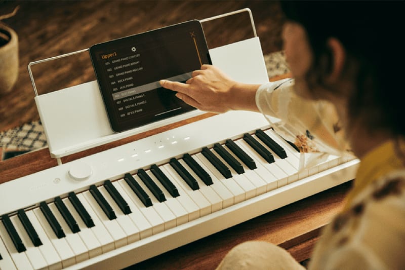 Una persona toca una tableta colocada en el soporte transparente para partituras de un piano digital blanco para utilizar la aplicación Casio Music Space.