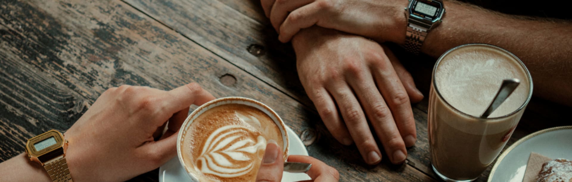 Vista desde arriba de cuatro manos sobre la mesa de una cafetería con diferentes tipos de café, una persona lleva el reloj de pulsera plateado ABL-100 y la otra, el dorado.