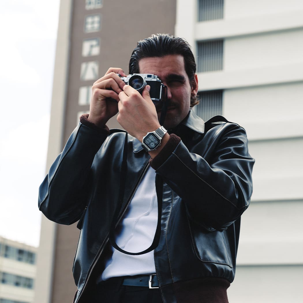 Un hombre delante de un rascacielos con una chaqueta de cuero negra, una camisa blanca y el AE-1200WHD-1A plateado de CASIO, mirando a través del objetivo de una cámara réflex.