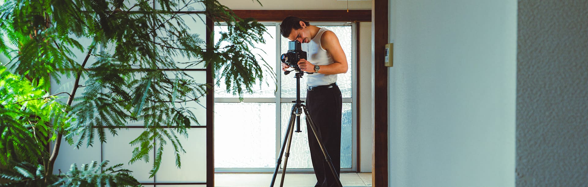 Un hombre con pantalones negros y camisa blanca lleva el reloj digital AE-1200WHD-1A en color plateado, de pie frente a un espejo y mirando a través del objetivo de su cámara fotográfica.