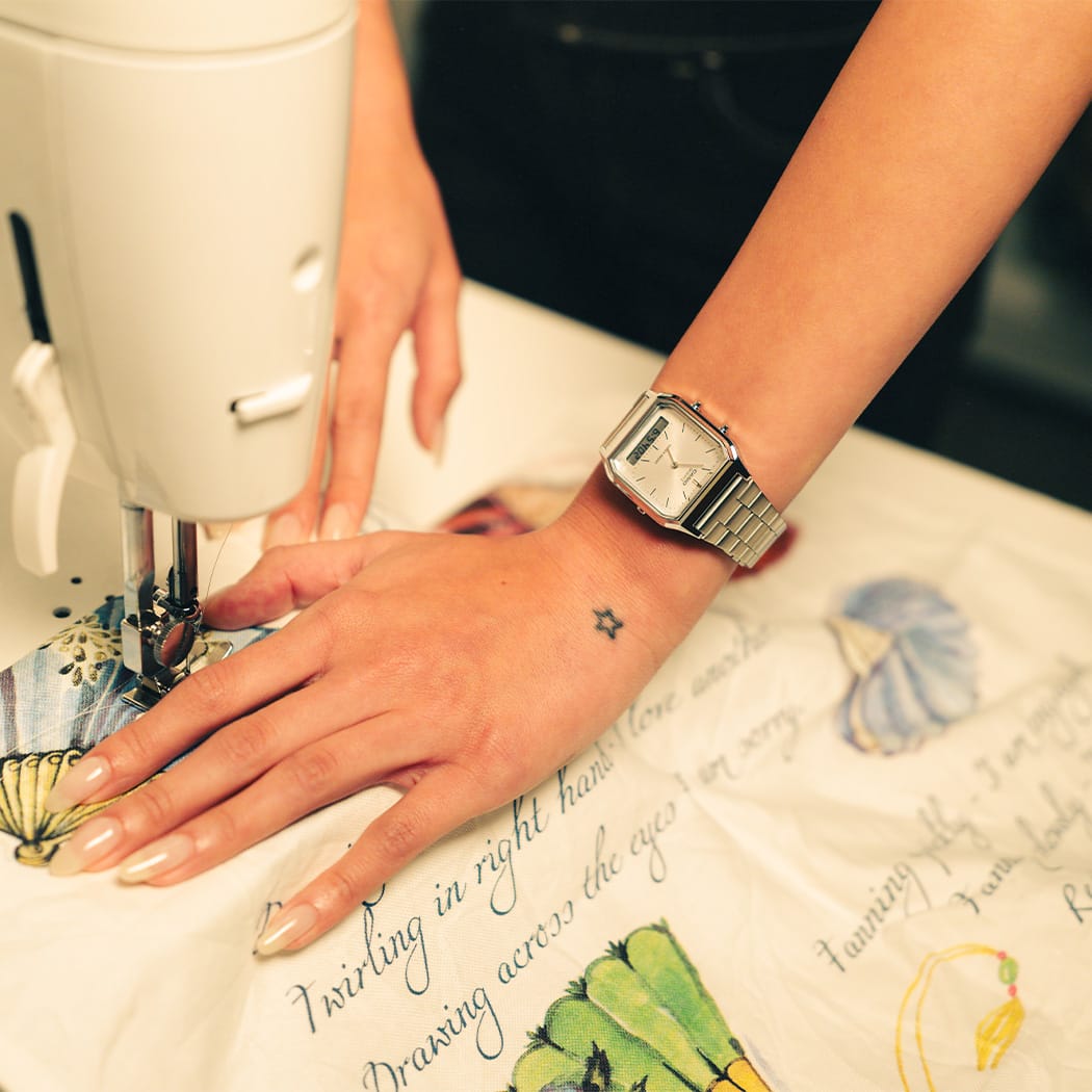 Primer plano de una mujer en una máquina de coser, con el reloj CASIO Vintage Collection "AQ-230A-7A" de color plateado con pantalla analógica en la muñeca.