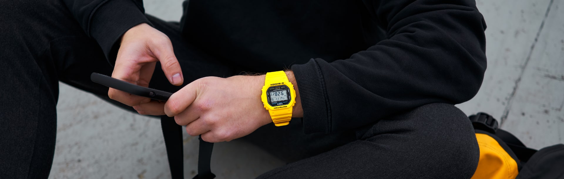 Una persona vestida de negro sostiene un smartphone y lleva en la muñeca un reloj G-SHOCK "DW-5600" de color amarillo