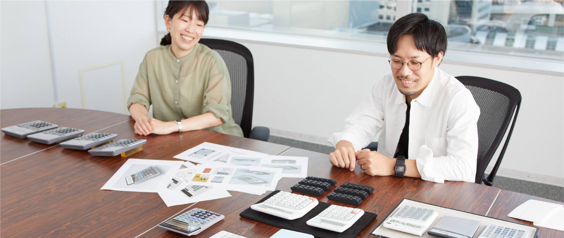 Product developers Kumira and Oka sit at a conference table with several prototypes of the ergonomic calculator on it