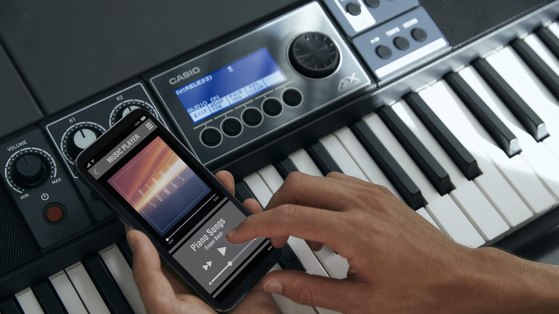 A man at the keyboard holding a mobile phone with a music player.