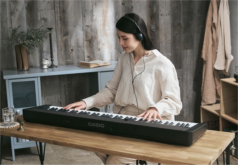 A young woman wears headphones and plays the black Casiotone CT-S1-76 digital piano