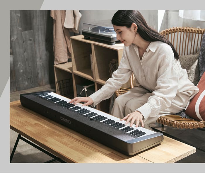 A woman sits in a wicker chair and leans forward to play the black Casiotone CT-S1-76 on the living room table in front of her