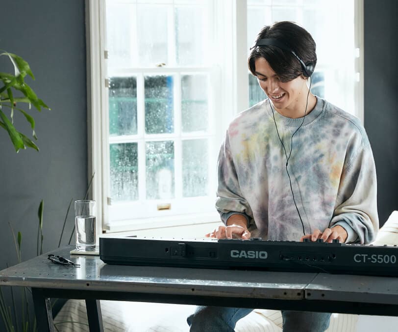 A young man is wearing headphones and smiling as he plays a Casiotone CT-S500