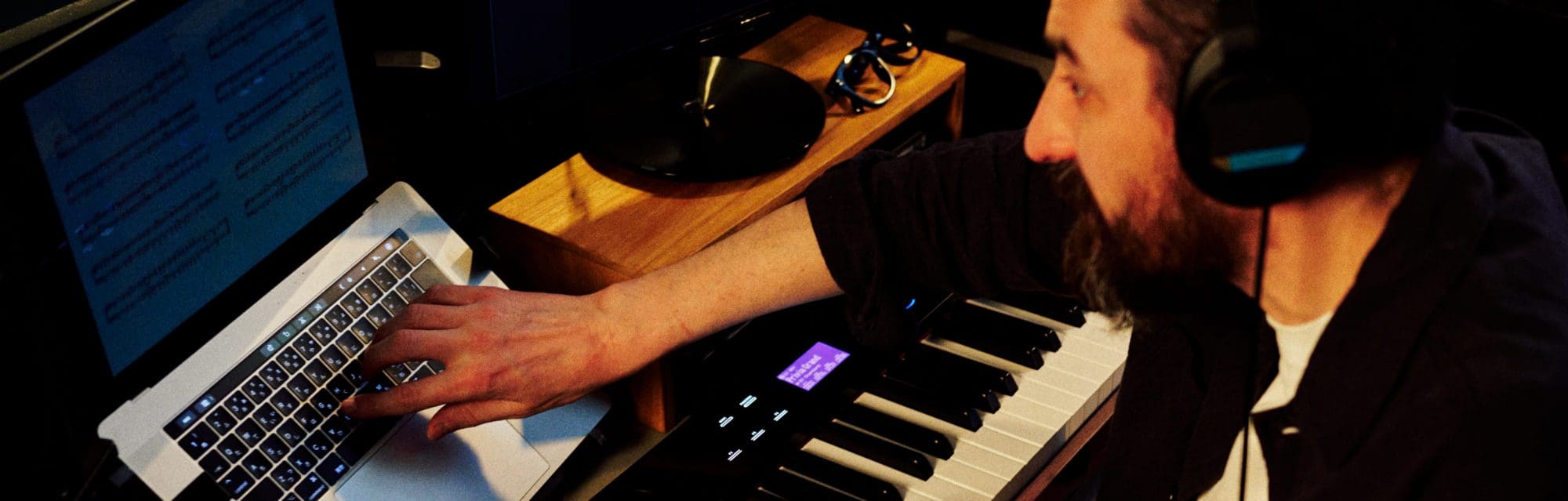 A man wearing headphones sits at the Privia PX-S6000 and types something into his laptop.