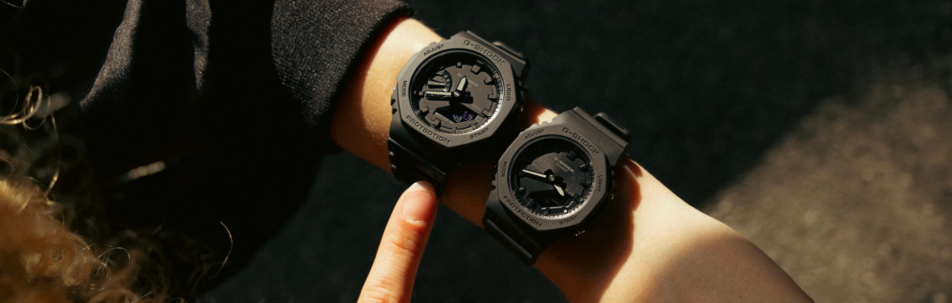 The larger black GA-2100-1A1 men's watch and the smaller GMA-P2100BB-1A ladies' watch, also in black, worn side by side on a slender wrist.