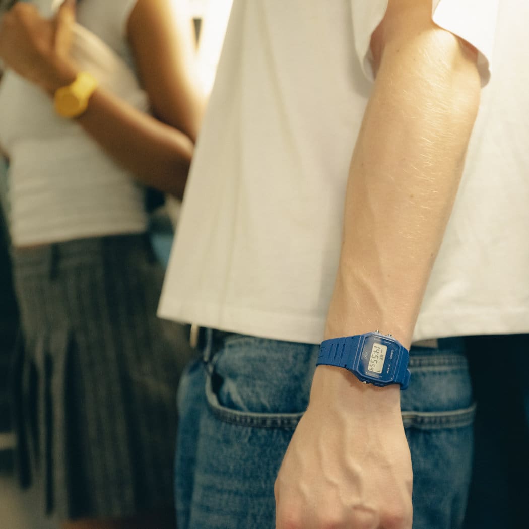 The blue F-91WB-2AEF on the wrist of a male model; a woman wearing the yellow MQ-24 can be seen in the background.