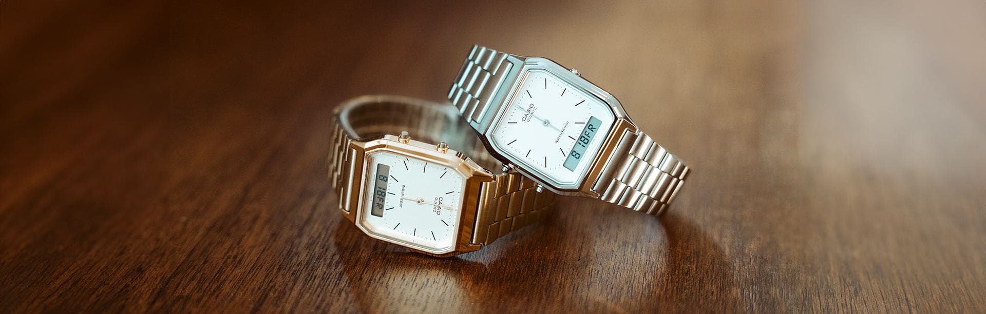 Two CASIO vintage watches in gold and silver lie on top of each other on a wooden table.
