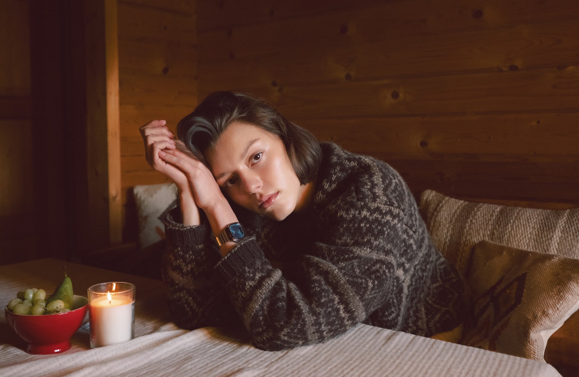 A woman in a wood cabin leaning onto a table with a white cloth lookind into the camera and wearing a woolen pullover and the AQ-240E-3A.