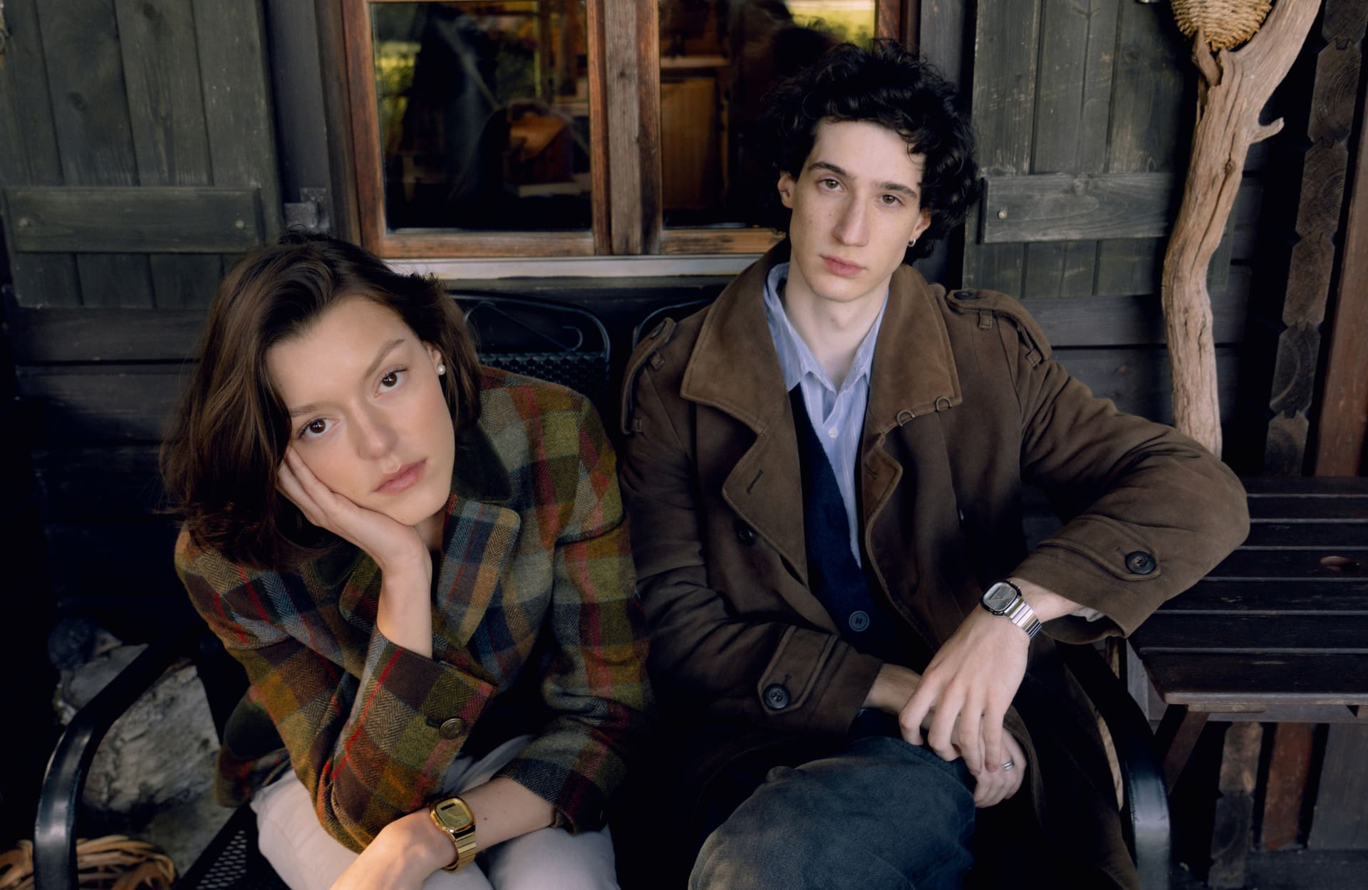 A couple sitting in front of a wooden cabin on a bench wearing brown-colored clothes and the CASIO Vintage Collection watches AQ-240E