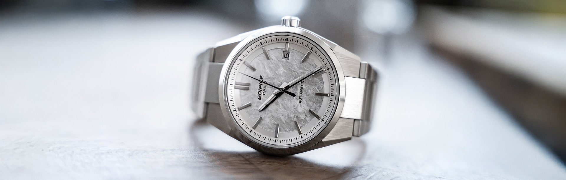 The EDIFICE EFK-110 automatic watch in silver with a white patterned analogue dial lies on its side on a light-coloured plate.