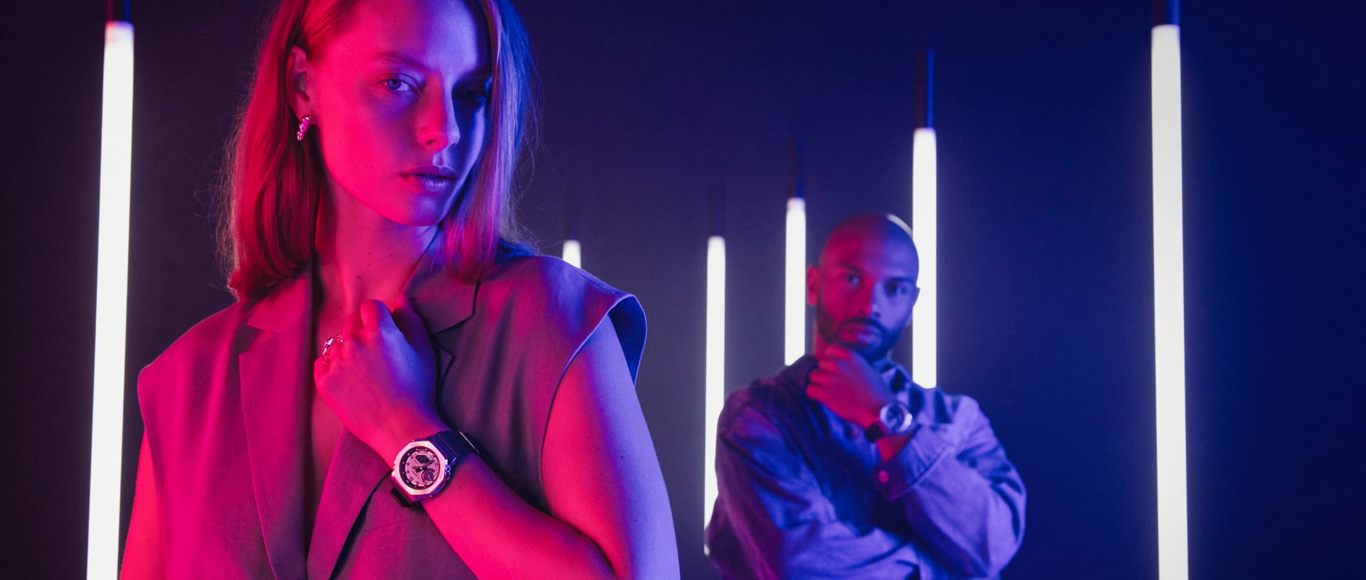 A woman stands in the foreground, holding up her arm, which is wearing the G-SHOCK GM-2100 watch with a metal bezel. In the background stands a man wearing the same model on his wrist.