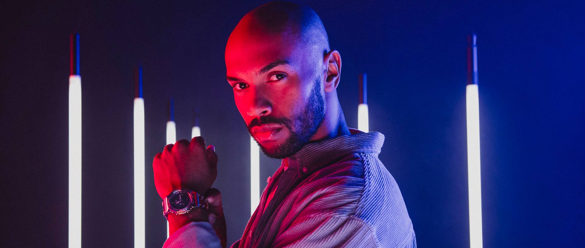Im Hintergrund sind längliche LED-Leuchten vertikal aufgehängt, Im Vordergrund ist ein Mann mit der G-SHOCK full-metal Uhr GM-B2100 in gold zu sehen.