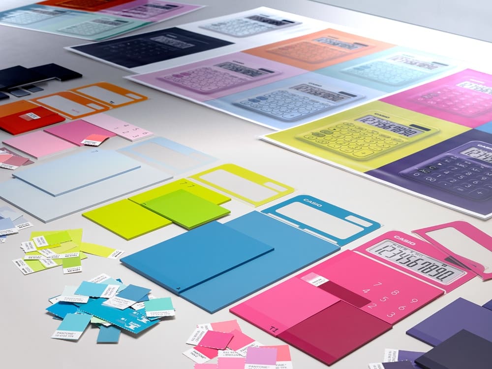 Différents matériaux et motifs colorés sont étalés sur une table, notamment des nuanciers et des modèles de design pour calculatrices.